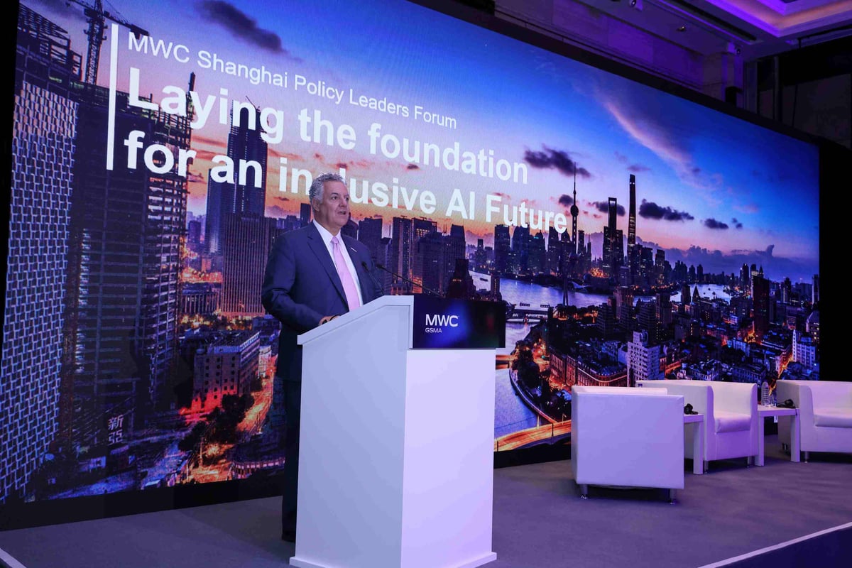A speaker stands at a podium on stage during the MWC Shanghai Policy Leaders Forum. Behind them, a large screen displays the event theme: “Laying the foundation for an inclusive AI Future!” The backdrop features a vibrant city skyline at sunset, with tall buildings silhouetted against colorful clouds. To the right of the podium, three white chairs are arranged, suggesting a panel discussion setup.