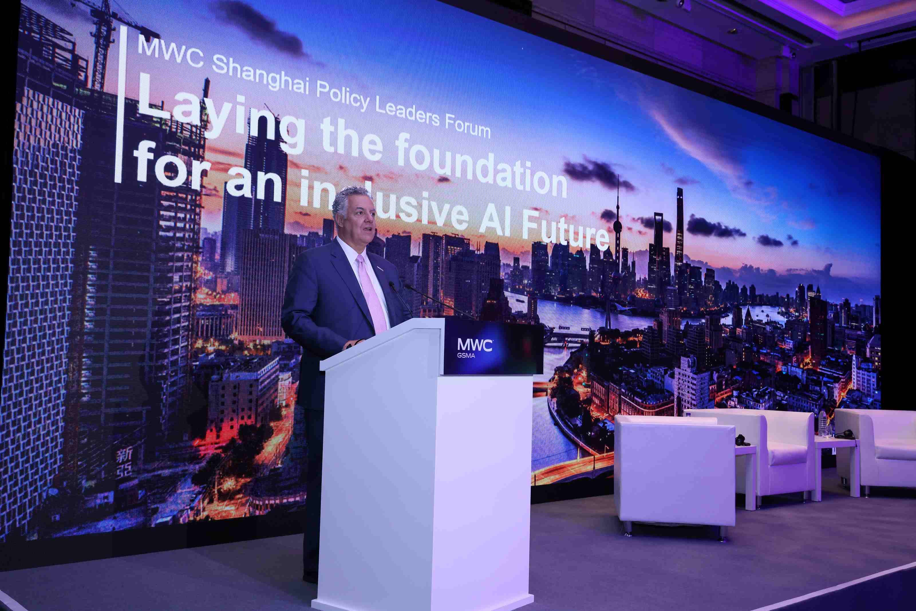 A speaker stands at a podium on stage during the MWC Shanghai Policy Leaders Forum. Behind them, a large screen displays the event theme: “Laying the foundation for an inclusive AI Future!” The backdrop features a vibrant city skyline at sunset, with tall buildings silhouetted against colorful clouds. To the right of the podium, three white chairs are arranged, suggesting a panel discussion setup.